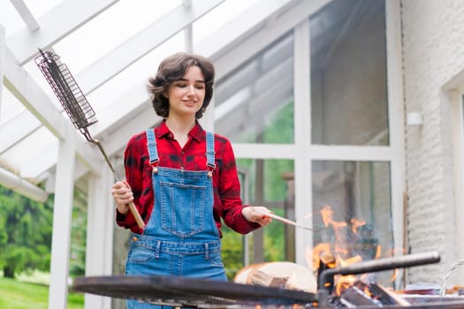 Barbeque party in garden with young woman in denim overalls and red plaid shirt in a country house on the terrace preparing meat skewers