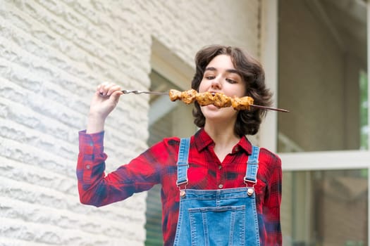 Barbeque party in garden with young woman in denim overalls and red plaid shirt in a country house on the terrace preparing meat skewers