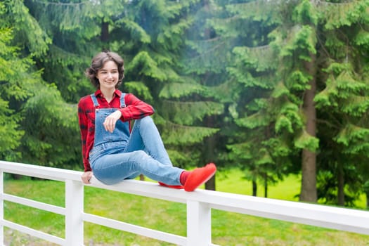 Happy young woman in blue denim overalls and red shirt sits on wooden fence on the terrace in the backyard, against the backdrop of a green lawn and forest on a sunny day