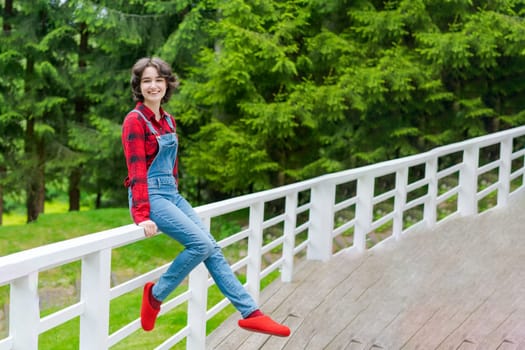 Happy young woman in blue denim overalls and red shirt sits on wooden fence on the terrace in the backyard, against the backdrop of a green lawn and forest on a sunny day