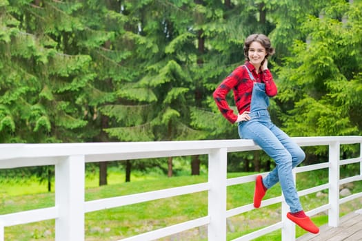 Happy young woman in blue denim overalls and red shirt sits on wooden fence on the terrace in the backyard, against the backdrop of a green lawn and forest on a sunny day