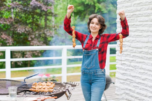 Barbeque party in garden with young woman in denim overalls and red plaid shirt in a country house on the terrace preparing meat skewers