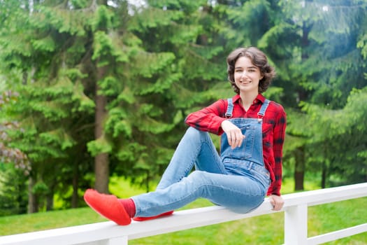 Happy young woman in blue denim overalls and red shirt sits on wooden fence on the terrace in the backyard, against the backdrop of a green lawn and forest on a sunny day