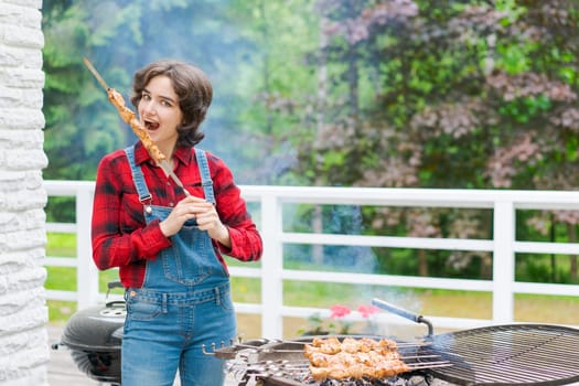 Barbeque party in garden with young woman in denim overalls and red plaid shirt in a country house on the terrace preparing meat skewers