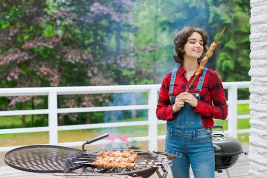 Barbeque party in garden with young woman in denim overalls and red plaid shirt in a country house on the terrace preparing meat skewers