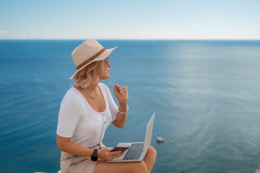 Freelance women sea working on the computer. Good looking middle aged woman typing on a laptop keyboard outdoors with a beautiful sea view. The concept of remote work