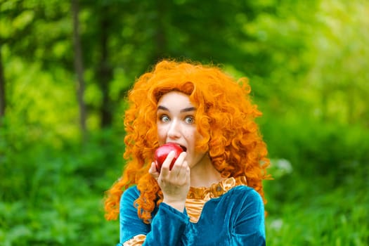 Curly red-haired girl fairy in nature with long curly hair in a blue dress eats a big red apple