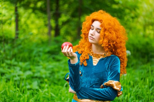 Curly red-haired girl fairy in nature with long curly hair in a blue dress eats a big red apple