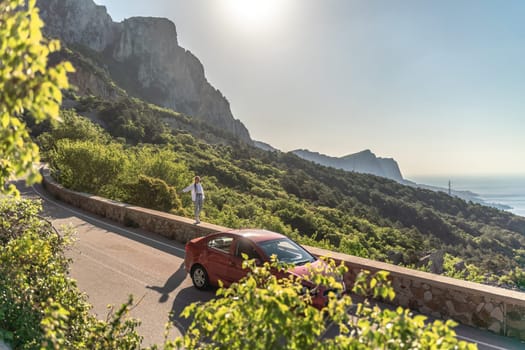 Mountain road woman. She walks along a parapet road in the middle of a mountainous area, a car is parked nearby. She is dressed in jeans and a white shirt, her hair is braided