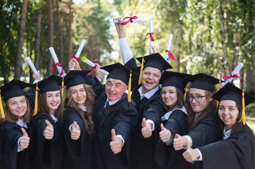 A group of graduates in robes give a thumbs up outdoors. Elderly student