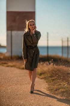 Portrait blonde sea cape. A calm young blonde in a khaki raincoat stands on the seashore against the backdrop of a lighthouse