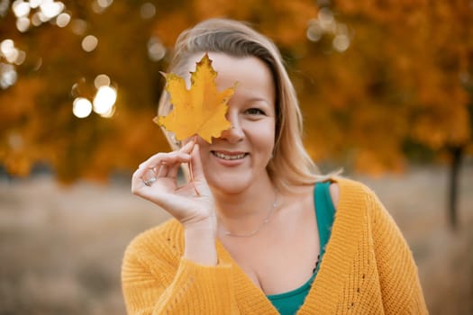 an autumn woman in a green dress holds an autumn leaf in front of her face, against the background of an autumn tree.