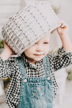 Little girl 1 years old with a basket on her head. Funny child plays puts a basket on her head like a hat and laughs, she is dressed in a plaid shirt and denim overalls. Funny moments with kids