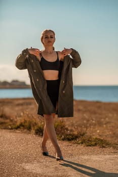 Portrait blonde sea cape. A calm young blonde in an unbuttoned khaki raincoat walks along the seashore, under a raincoat a black skirt and top.