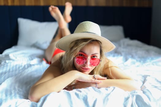 A young smiling woman in a red bikini stands near the bed in a cozy hotel room