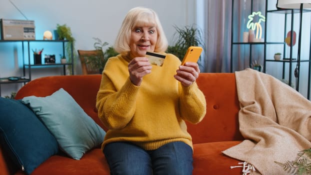 Grandmother senior using credit bank card and smartphone while transferring money, purchases online shopping cashless, order food delivery, pay bills at home apartment indoor. Elderly woman on couch