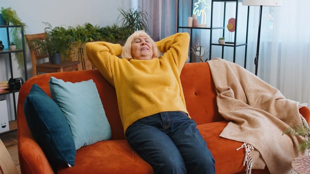 Tired mature grandmother woman enjoy relaxing on home sofa in room. Happy senior pensioner sits on couch resting napping after hard working day, closed his eyes, put her hands behind head, stress free