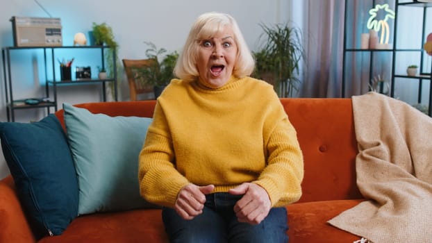 Excited senior grandmother woman on couch living room watching interesting TV serial sport game, film online social media movie content at home apartment. Elderly old pensioner enjoying entertainment