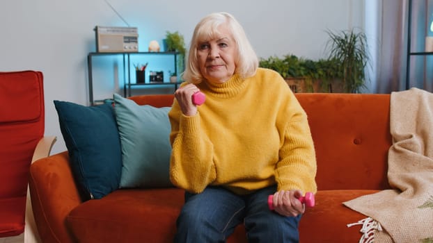 Senior grandmother woman doing dumbbells weight lifting fitness exercises at home apartment indoors. Elderly pensioner workout training strength practicing effort in room on couch. Sport motivation