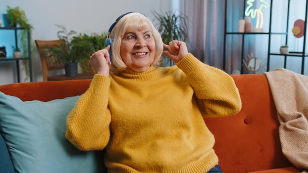 Happy overjoyed elderly grandmother woman in wireless headphones relaxing sits on couch at home apartment choosing listening favorite energetic disco dancing music. Weekend daytime leisure activities