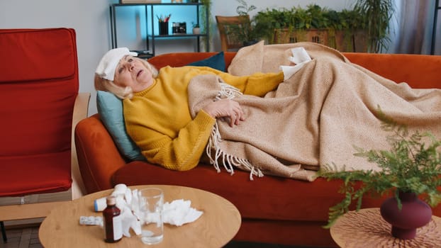 Grandmother ill woman suffering from cold or allergy lying on sofa in room at home apartment. Sick senior lady with towel on head blows sneezes wipes snot into napkin. Coronavirus quarantine, pandemic