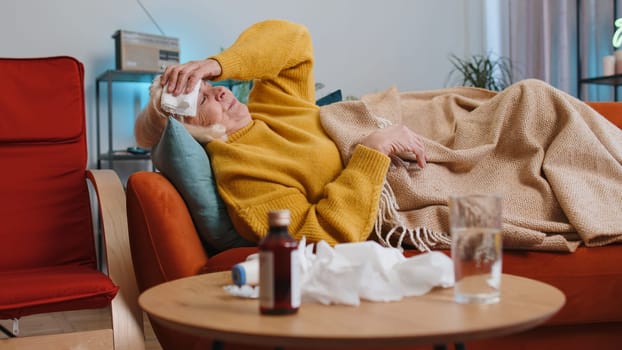 Grandmother ill suffering from cold or allergy pain lying on couch in living room at home. Sick mature woman with towel on head blows sneezes wipes snot into napkin. Coronavirus quarantine pandemic