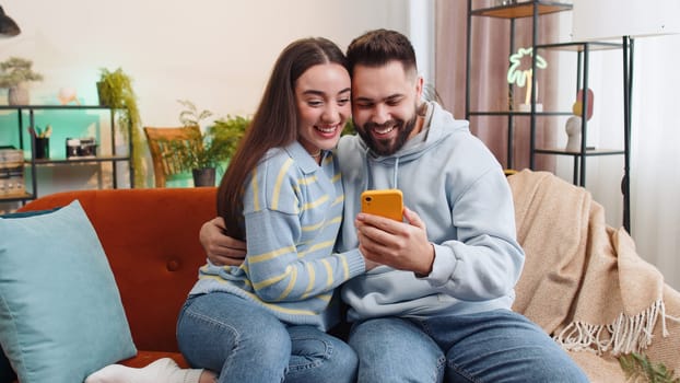 Family couple man woman use mobile smartphone found out great big win good news celebrate lottery jackpot lucky victory, playing game shopping online at home. Husband and wife together on sofa in room