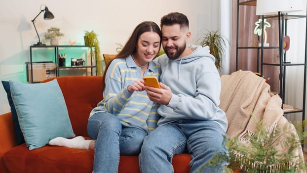 Family married couple use mobile smartphone found out great big win good news celebrate lottery jackpot lucky victory, playing game, shopping online at home. Husband and wife together on sofa in room