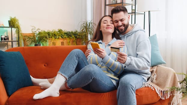 Family married couple man woman using credit bank card and smartphone while transferring money, purchases online shopping, order food delivery at home. Husband and wife together on sofa in living room