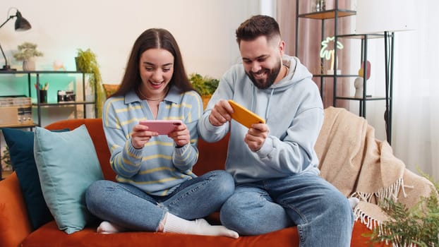 Happy family married couple man woman enthusiastically playing racing or shooter video app games online on smartphone at home. Husband and wife cheating together while playing on sofa in living room