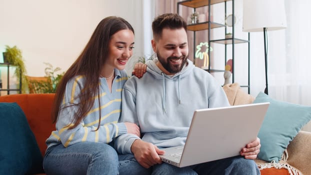 Young family marriage couple man woman making video laptop webcam conference call with friends or family, enjoying pleasant conversation at home. Husband wife laughing, waving hello on sofa in room