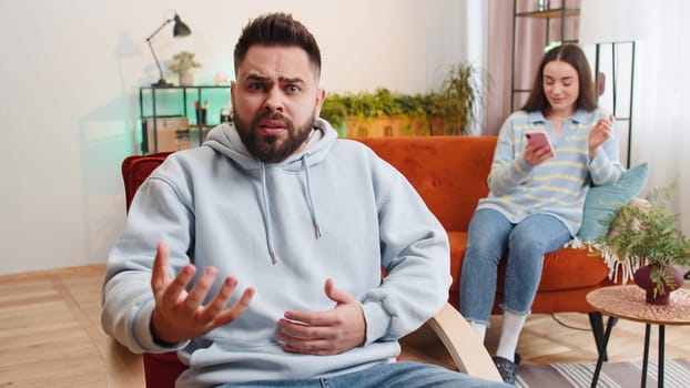 Unhappy man looking at camera, quarreling with woman addicted on online shopping wasting money with credit card, smartphone. Conflict problem, crisis in married family couple. Husband and wife at home