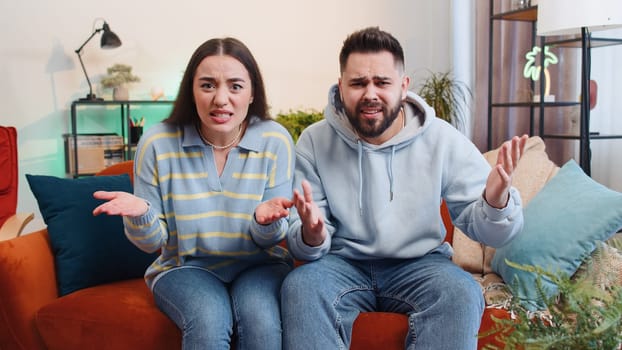 What. Why. Young family married couple man woman raising hands in indignant expression, asking reason of failure, demonstrating disbelief irritation by troubles at home. Husband wife together on sofa