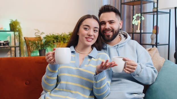 Young smiling family married couple man woman drinking a cup of warm coffee or herbal tea at home apartment in the morning before going to work. Husband and wife together enjoying comfortable relaxing