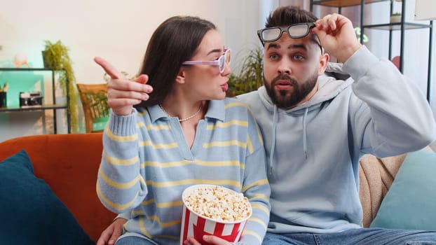 Excited young family marriage couple man woman eating popcorn and watching interesting tv serial, sport game, film, online social media movie content at home. Husband and wife together on sofa in room