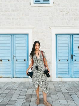 Girl photographer with digital cameras on straps stands near an old stone house. High quality photo