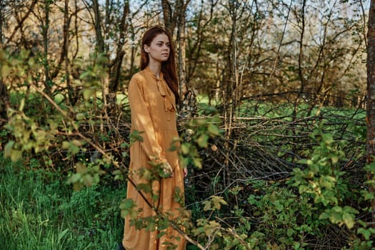 a woman with long hair walks in the shade near the trees, dressed in a long orange dress, enjoying the weather and the weekend, inspecting the trees. The theme of privacy with nature. High quality photo