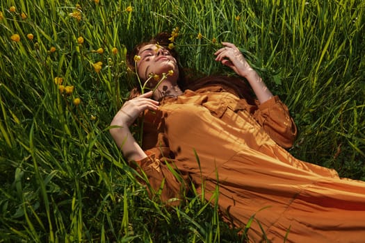 a calm woman with long red hair lies in a green field with yellow flowers, in an orange dress with her eyes closed, spreading her arms to the sides, enjoying peace and recuperating. High quality photo