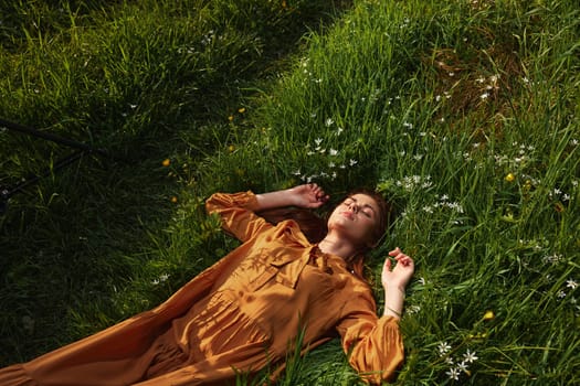 a happy, relaxed woman, resting lying in the green grass, in a long orange dress, with her eyes closed and a pleasant smile on her face, holding her hands near her face, enjoying harmony with nature and recuperating. High quality photo