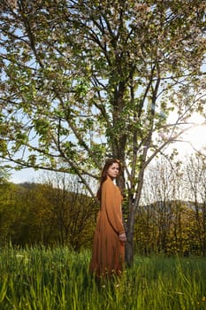 a sweet, attractive woman with long red hair stands in the countryside near a flowering tree in a long orange dress and looks at the camera with a slight smile on her face. High quality photo