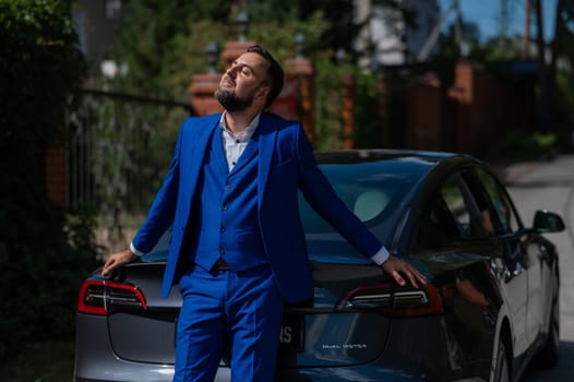 Caucasian bearded man in a blue suit stands near a black car in the countryside in summer