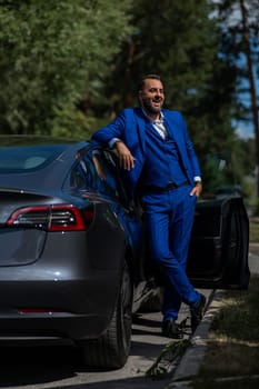 Caucasian bearded man in a blue suit gets out of a black electro car in the countryside in summer