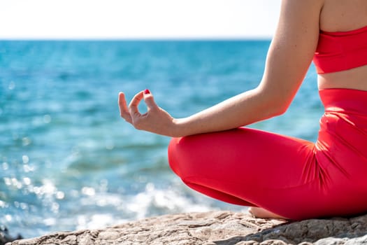 A young woman in red leggings and a red top with long loose hair practices yoga outdoors by the sea on a sunny day. Women's yoga, fitness, Pilates. The concept of a healthy lifestyle, harmony