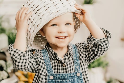 Little girl 1 years old with a basket on her head. Funny child plays puts a basket on her head like a hat and laughs, she is dressed in a plaid shirt and denim overalls. Funny moments with kids