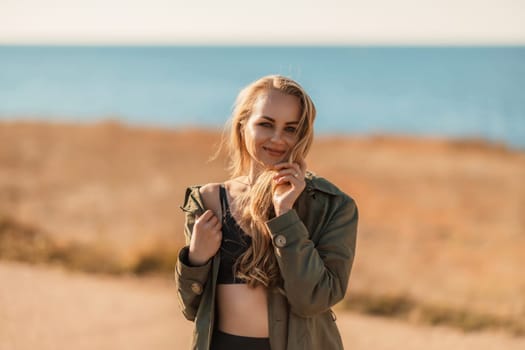 Portrait blonde sea cape. A calm young blonde in an unbuttoned khaki raincoat stands on the seashore, under the raincoat there is a black skirt and top.