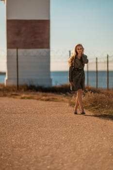 Portrait blonde sea cape. A calm young blonde in a khaki raincoat stands on the seashore against the backdrop of a lighthouse