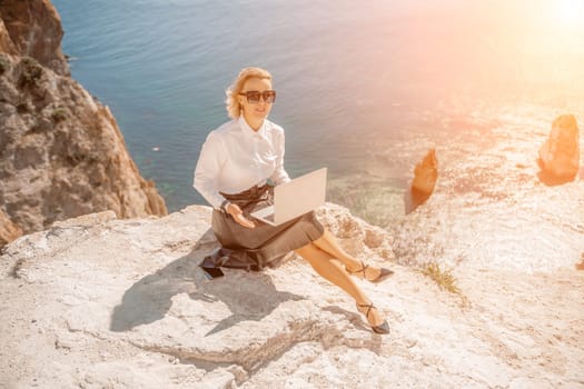 Business woman on nature in white shirt and black skirt. She works with an iPad in the open air with a beautiful view of the sea. The concept of remote work