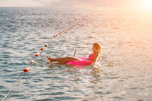 Woman works on laptop in sea. Freelancer, blond woman in sunglases floating on an inflatable big pink donut with a laptop in the sea at sunset. People summer vacation rest lifestyle concept