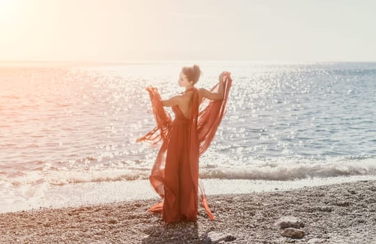 Side view a Young beautiful sensual woman in a red long dress posing on a rock high above the sea during sunrise. Girl on the nature on blue sky background. Fashion photo.