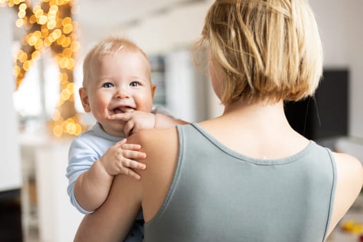 Blonde mother holding her adorable cheerful little infant baby boy. Cute infant child looking in camera and smiling at home